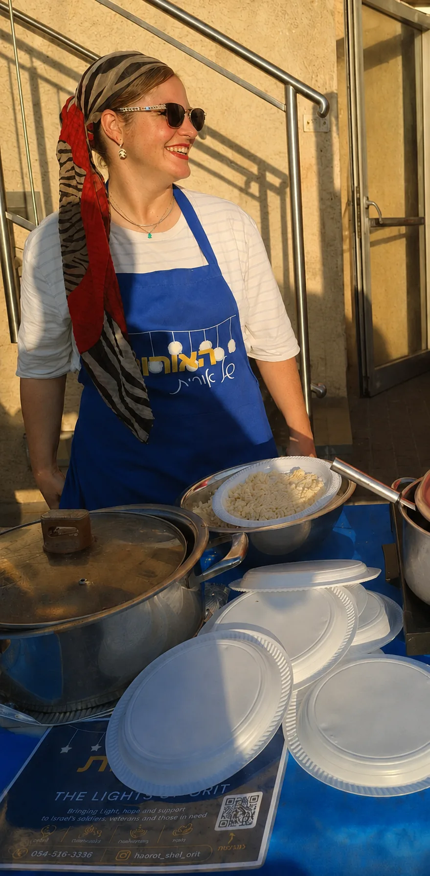 A smiling Lights of Orit volunteer in a branded apron serving homemade food at an evening of appreciation for soldiers